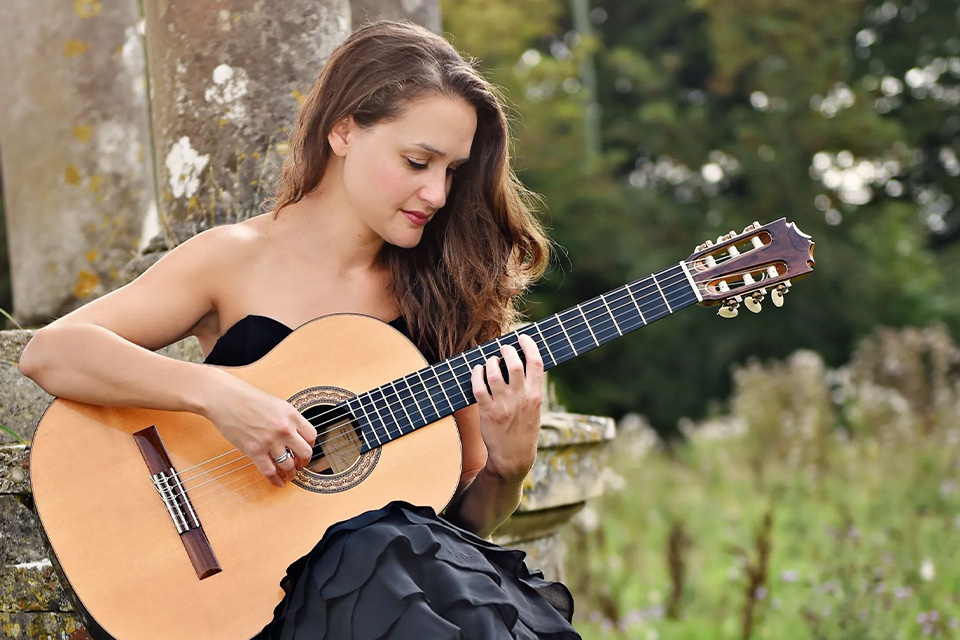 Amanda Cook wears a black dress a plays her guitar in a grassy field