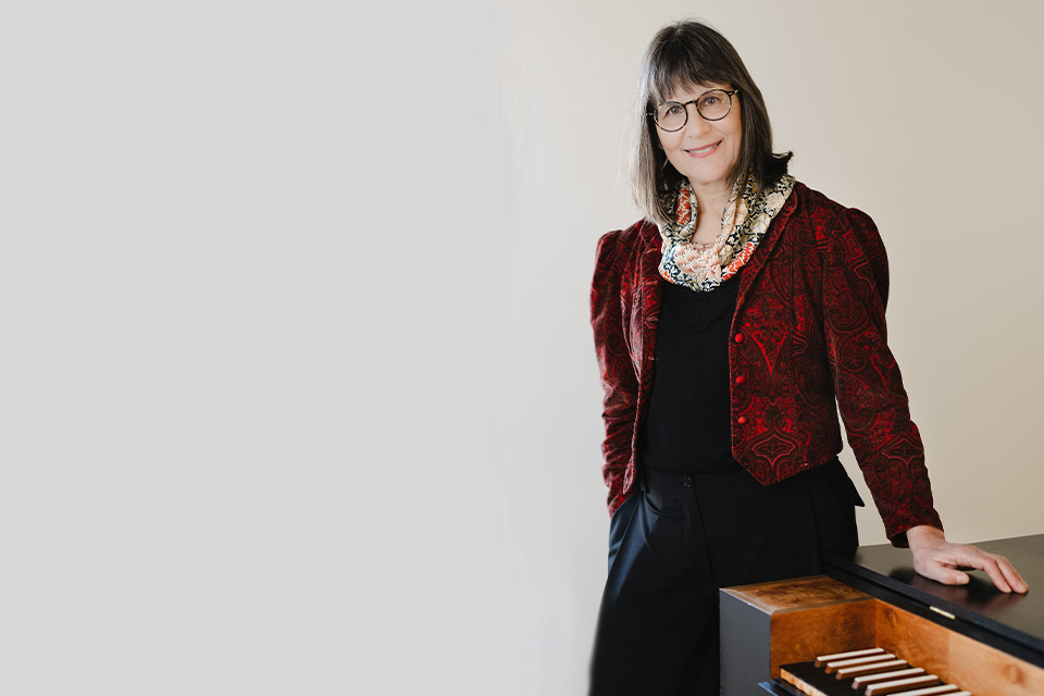 Marcia Hadjimarkos wears a dark red jacket and rests her hand on a clavichord