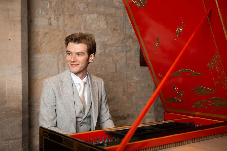 Nathaniel Mander wears a light grey suit and sits in front of a red harpsichord