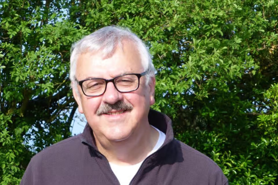 A man with grey hair and glasses stands in the sun outside in front of a leafy background