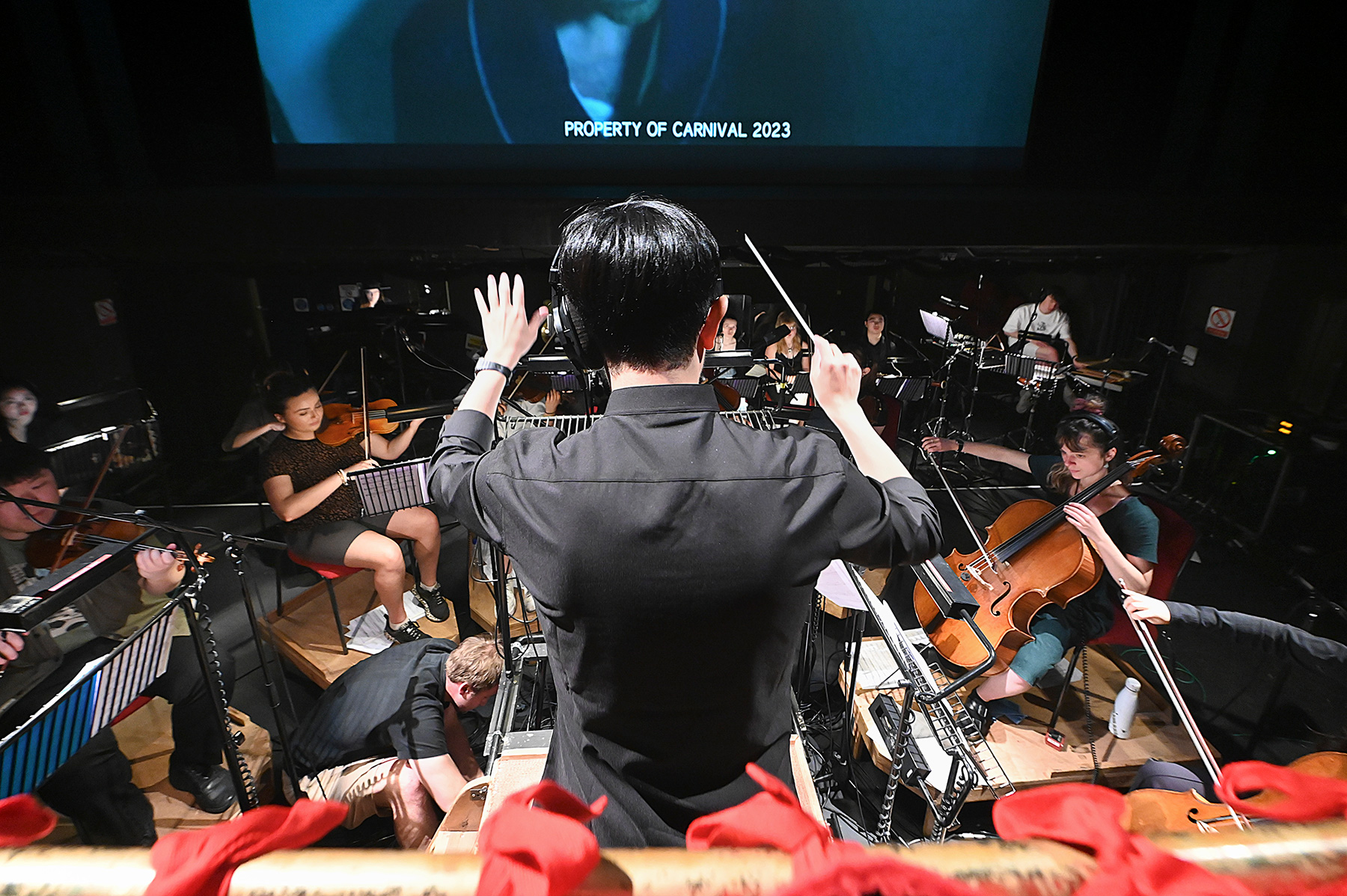 A male student leading an orchestra in the orchestra pit, with a screen on a stage above.