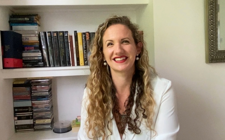 a woman with curly dark blonde hair, smiling, sitting against a bookshelf