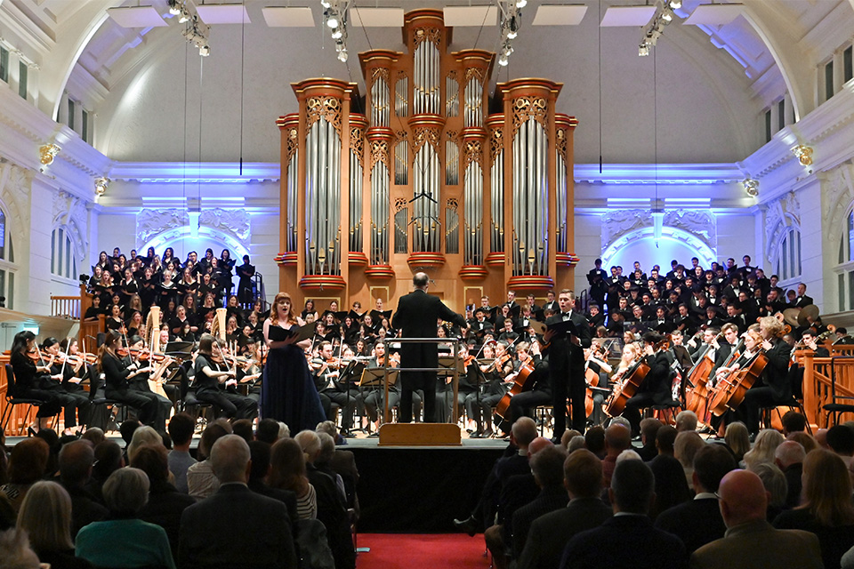 Martyn Brabbins conducts a large orchestra and chorus on a brightly lit stage, and audience listening attentively