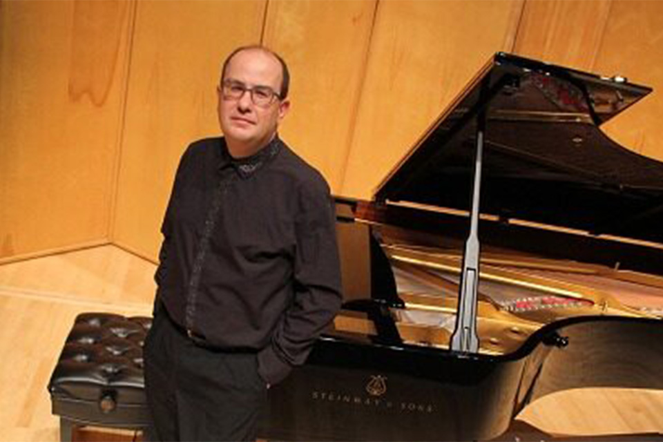 Alexandre Dossin leans against a piano on a brightly lit stage, wearing a black shirt and trousers with his hands in his pockets