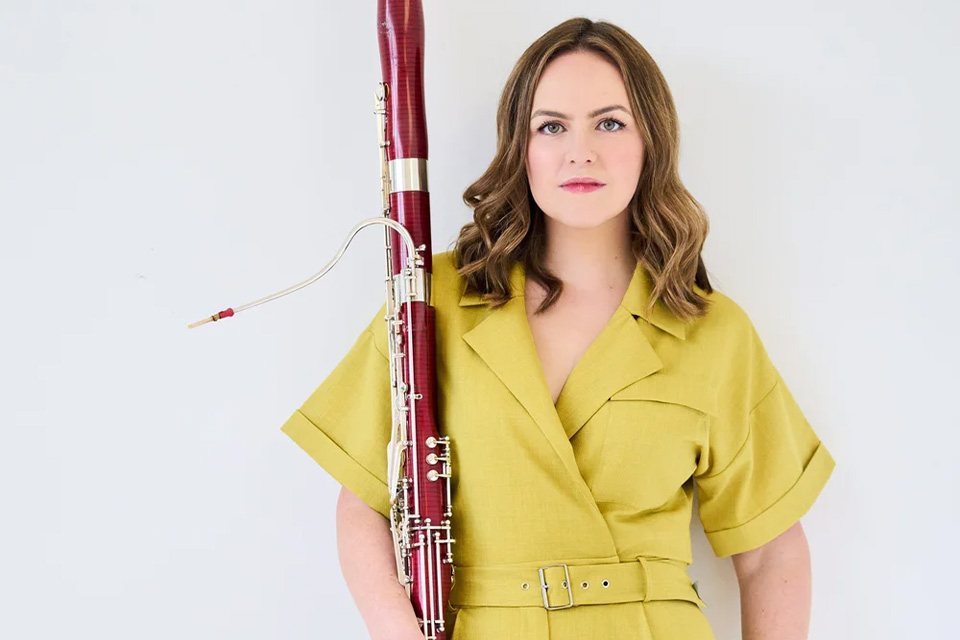 Amy Harman wears a yellow outfit whilst holding her bassoon and looking at the camera