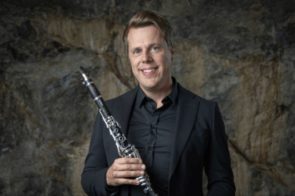 Andreas Sundén, wearing black, stands against a stone wall, smiling and holding his clarinet