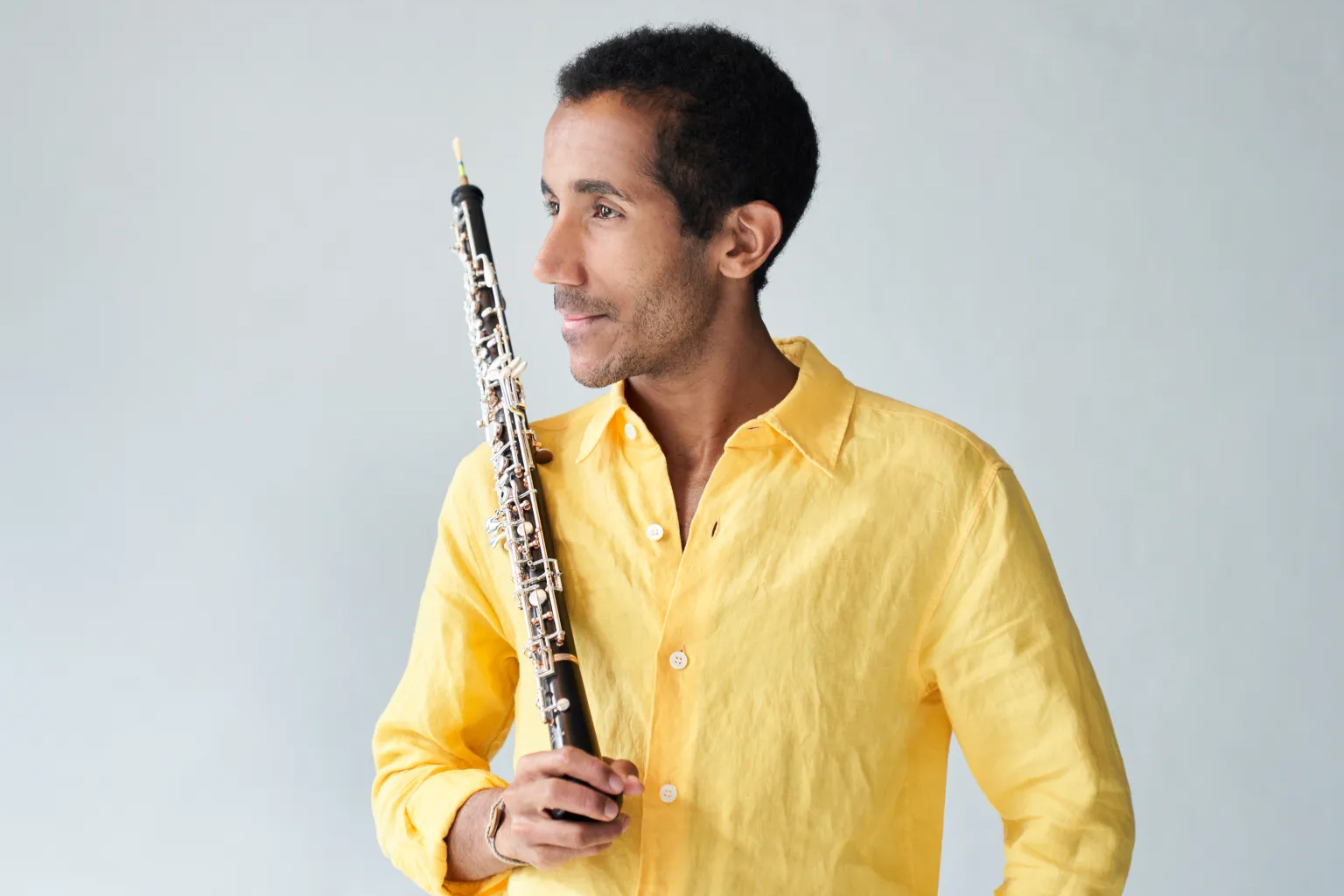 A man in a yellow shirt holds an oboe and looks off to the side