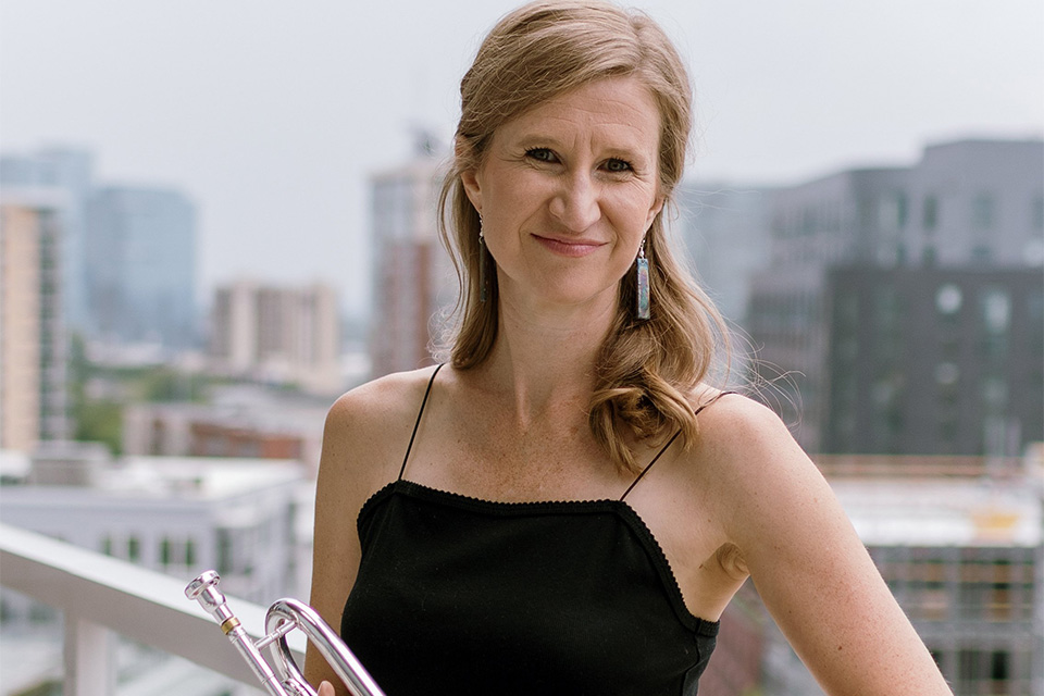 Ashley Hall-Tighe stands on a balcony with a cityscape behind her, holding her trimpet and smiling