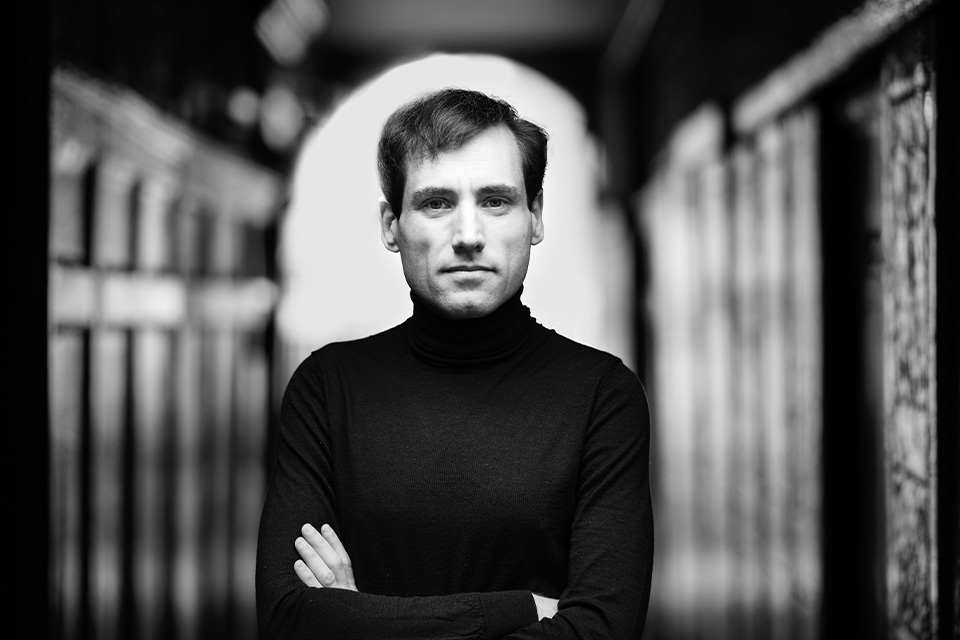 A black and white photo of Boris Giltburg standing in a corridor with his arms crossed, wearing a black turtleneck jumper