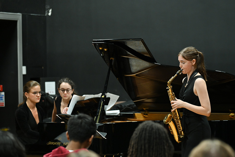 A young woman plays the saxophone to an audience, a piano accompanist behind her