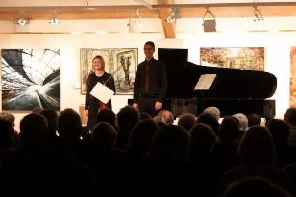 Two musicians stand on stage with a piano in front of an audience
