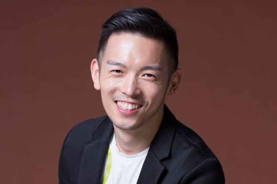 Chun-Chieh Yen wears a black blazer and white t-shirt against a dark red background, smiling