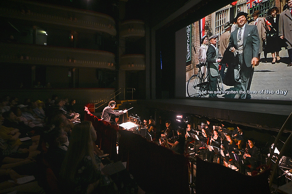 An orchestra performs in the pit of a theatre underneath a projection on screen