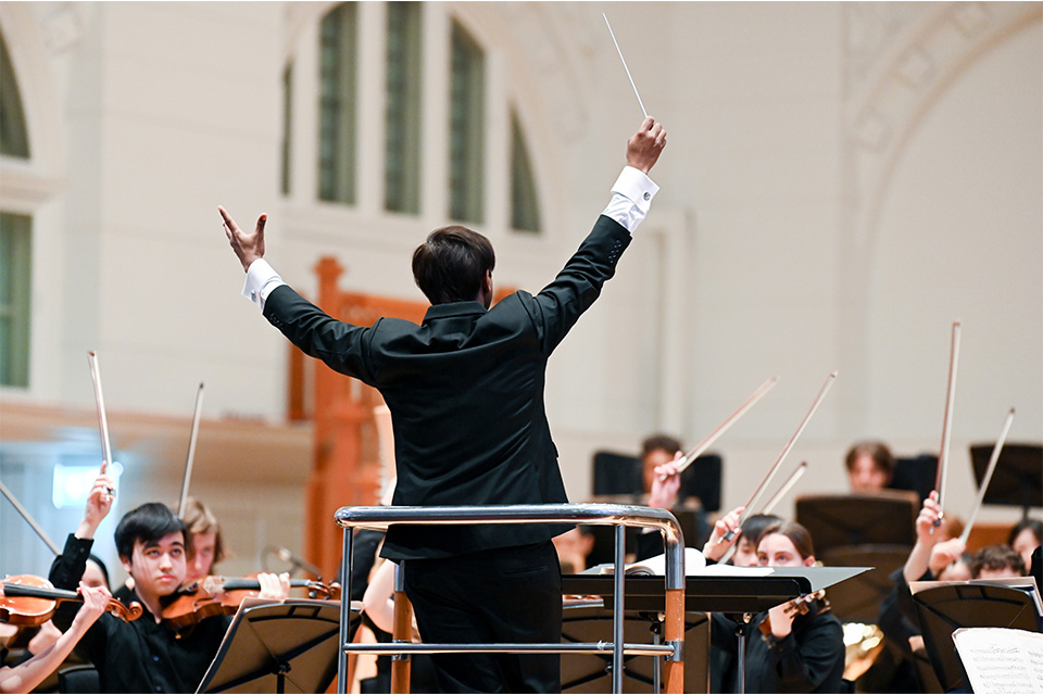 The silhouette of the back a conductor with his hands in the air, the orchestra in front of him with their bows in the air