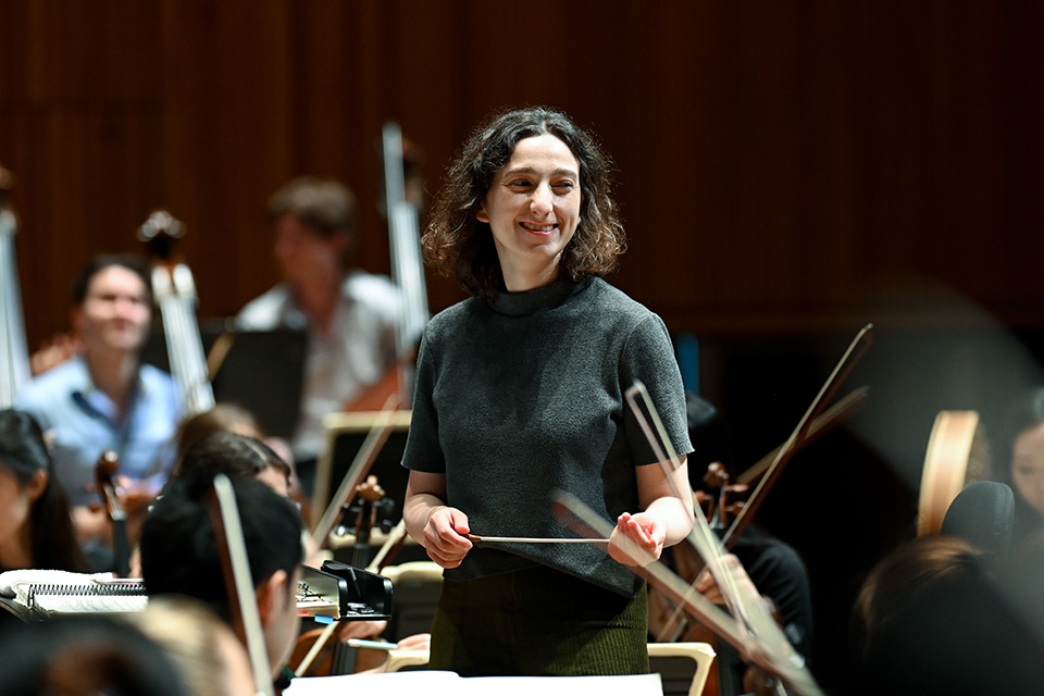 A woman stands on the conductors podium in front of an orchestra and smiles