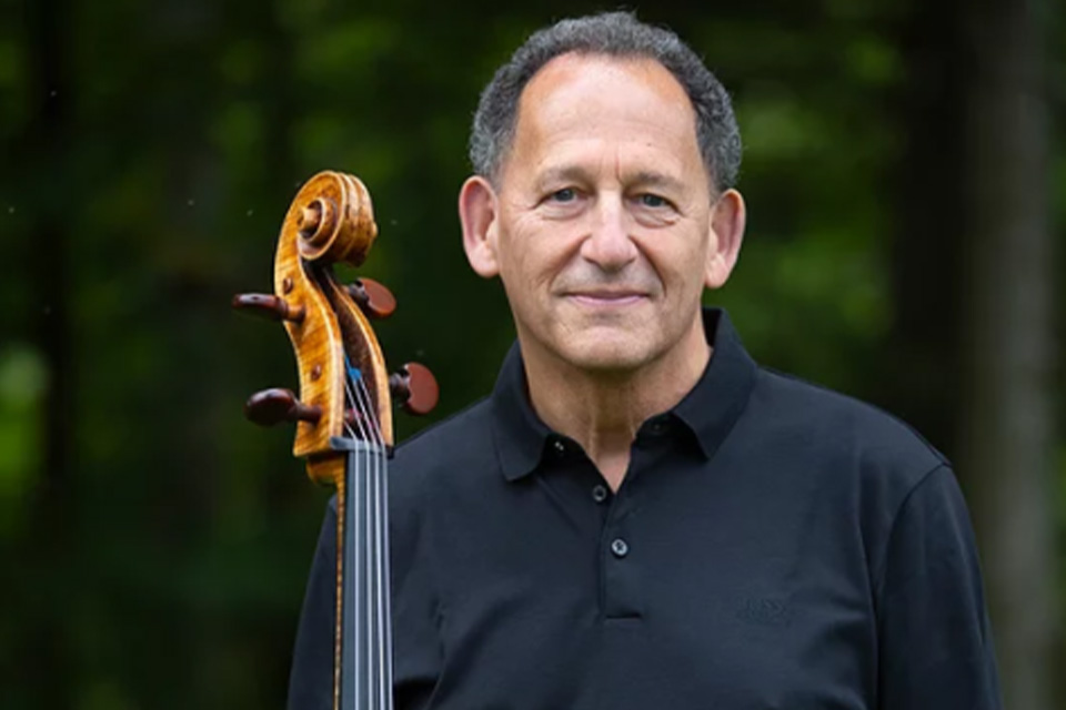 Denis Brott stands outside wearing a black shirt and holding his cello, a leafy background behind him