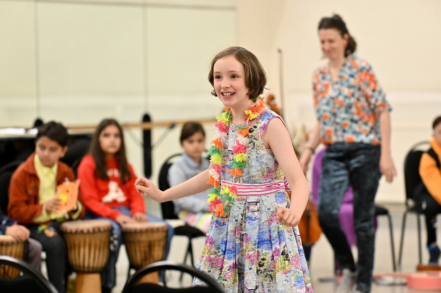A girl performs with other children playing percussion instruments and an adult dancing in the background