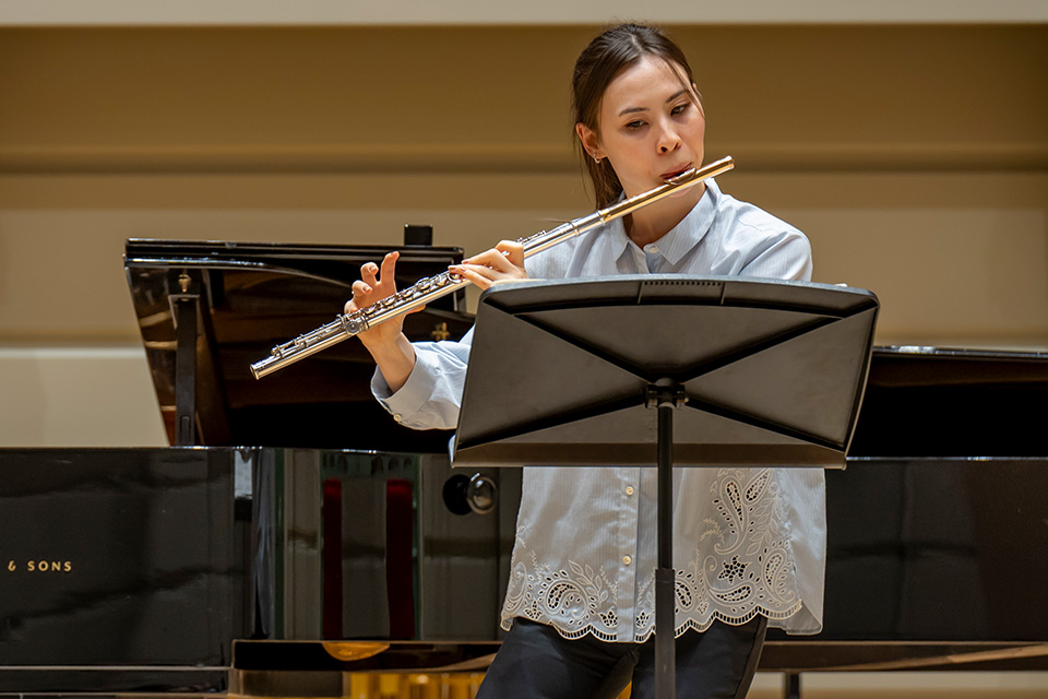A flautist wears a blue shirt and performs on stage in front of a piano