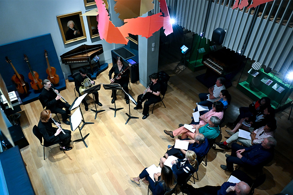 An aerial photo of a wind quintet performing in the RCM museum, while a small audience watches attentively