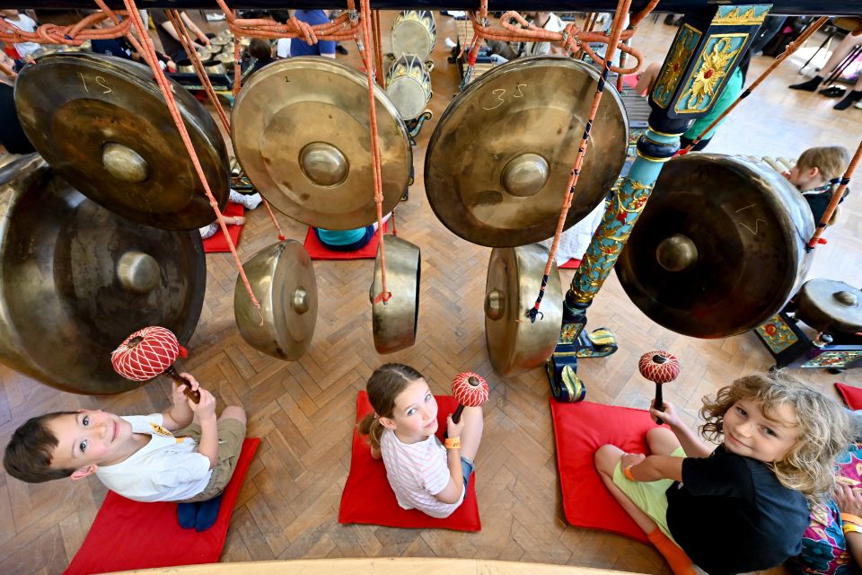 Three young children sit on the floor in front of large gongs, looking up at the camera