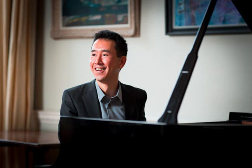 Hiro Takenouchi sits at the piano wearing a suit and smiles