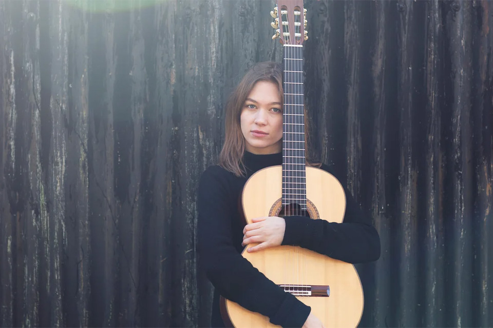 Hope Cramsie stands outside wearing a black top and holding her guitar