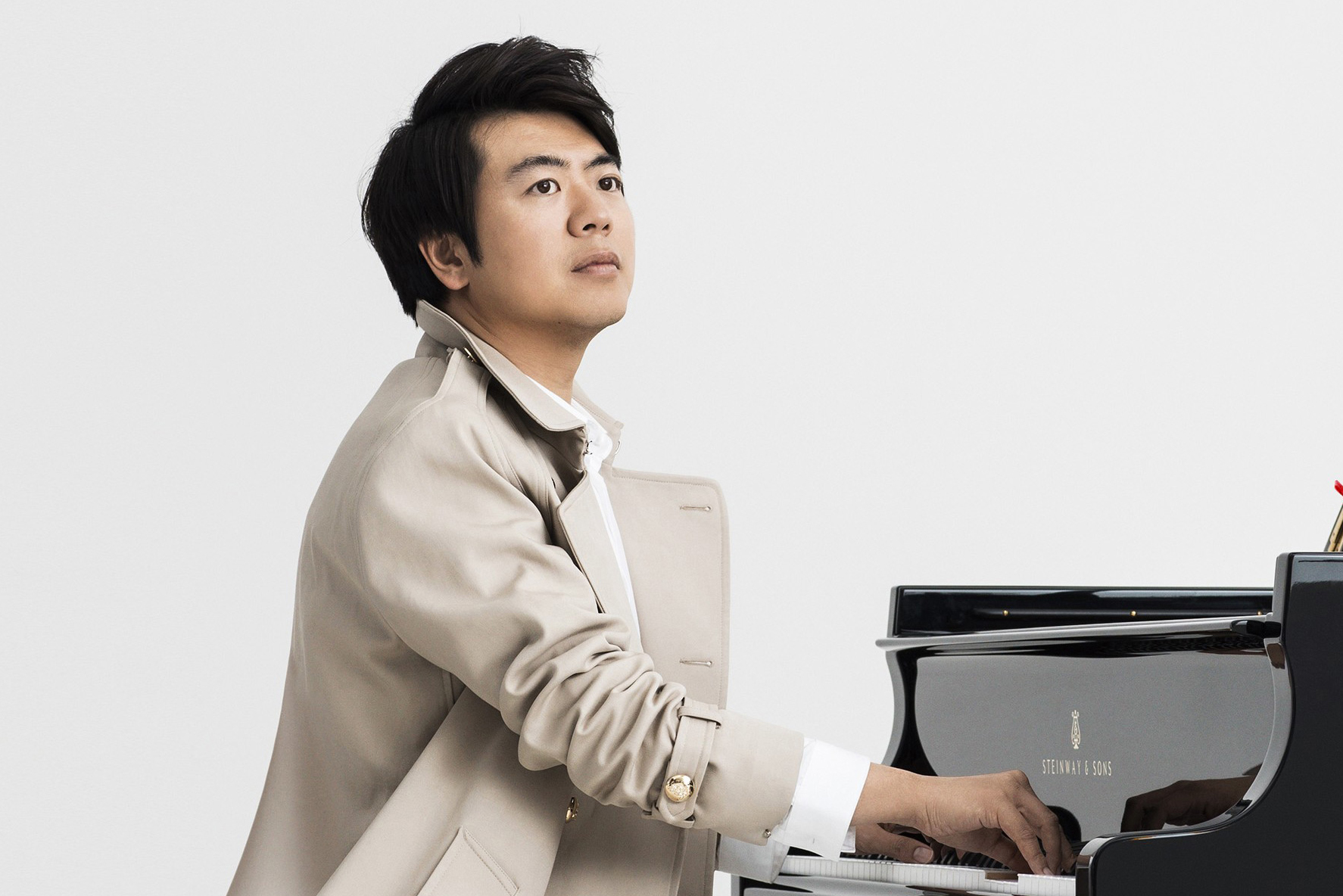 Lang Lang in a beige trenchcoat performing on a grand piano against a plain white background
