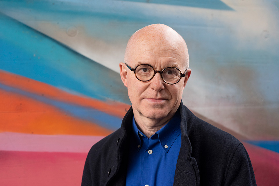 Laurence Cummings wears a blue shirt and stands outside with a multi-coloured wall behind him
