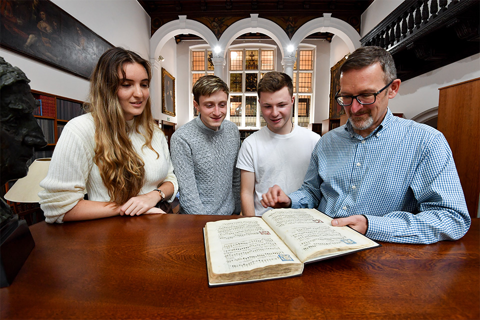 Four people stand around a table in the RCM library looking down at a large opera score