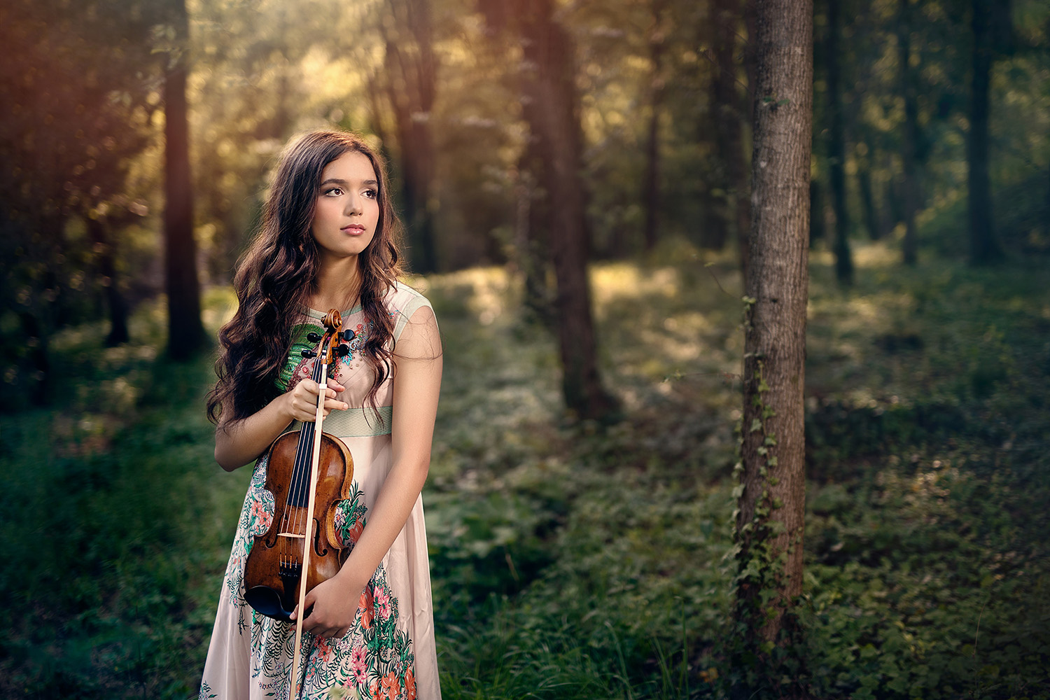 A woman with long dark hair holding a violin and standing in the woods