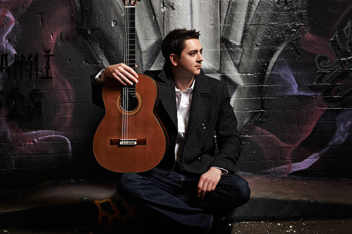 A man in a dark coat sitting cross-legged on the floor, looking off to the side and holding a guitar