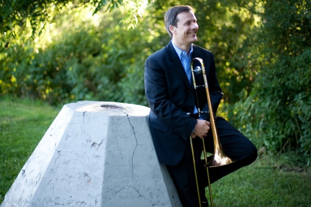 A man wearing a suit, holding a trombone and leaning against a stone structure in a park