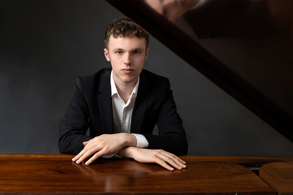 Matty Oxtoby wears a suit and blazer and leans on a piano with his hands folded over each other