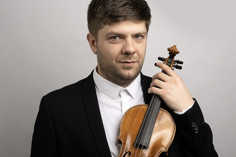 A man wearing a black suit and white shirt holding a violin and looking at the camera