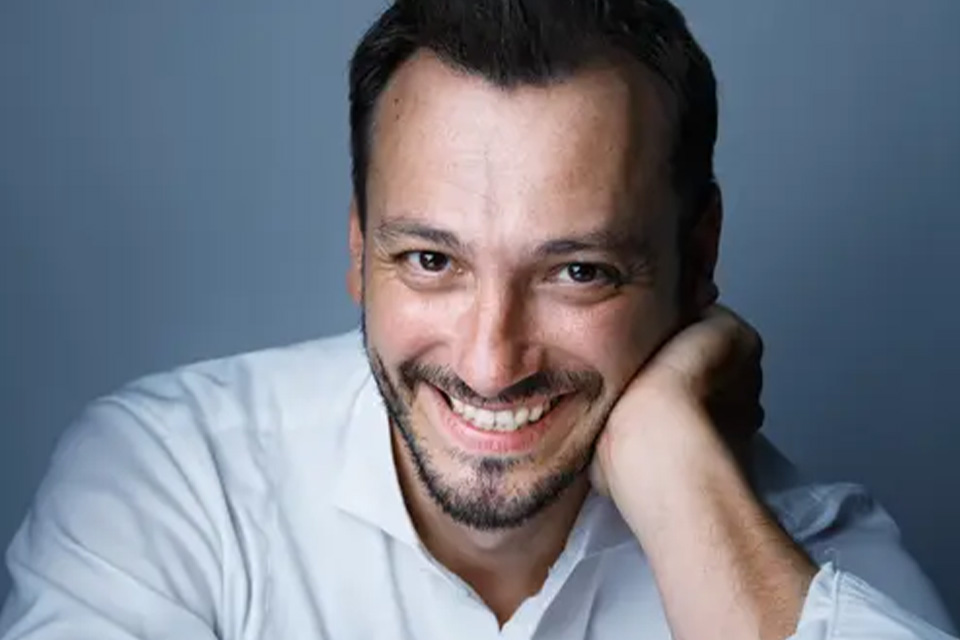 Maxim Rysanov rests his head on his hand and smiles against a blue background