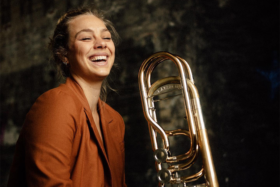 Maxine Troglauer, wearing a dark orange jacket, holds her trombone and laughs