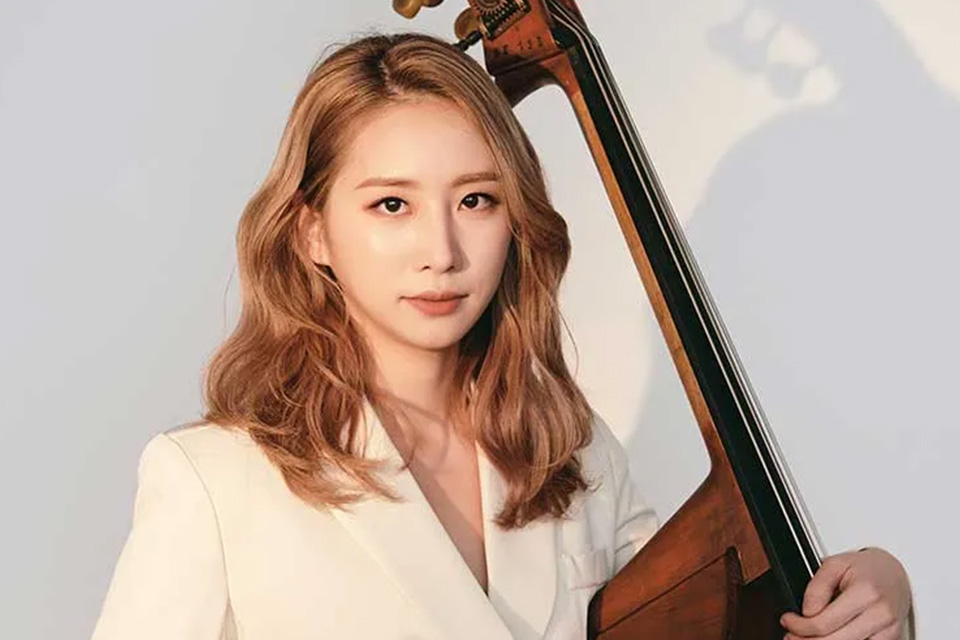 Mikyung Sung wears a white shirt and holds her double bass against a white background, her face lit up by a faint golden light