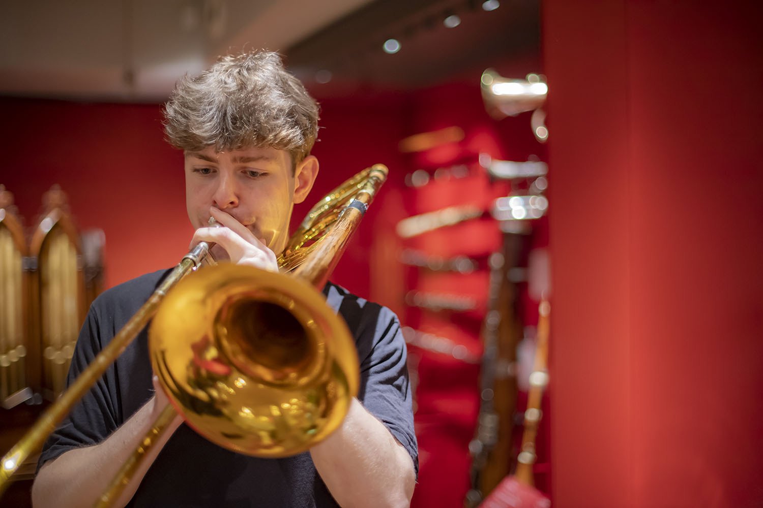 A man with short brown hear wearing a black t-shirt performs on a trombone in the Royal College of Music Museum