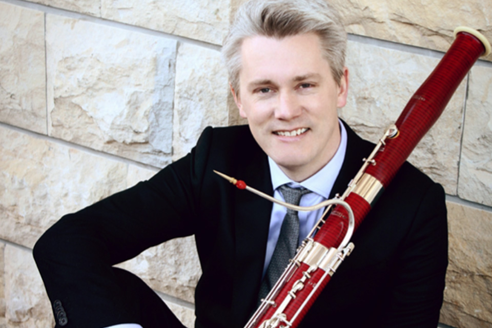 Ole Kristian Dahl sits against a wall in a suit and tie, holding his bassoon and smiling