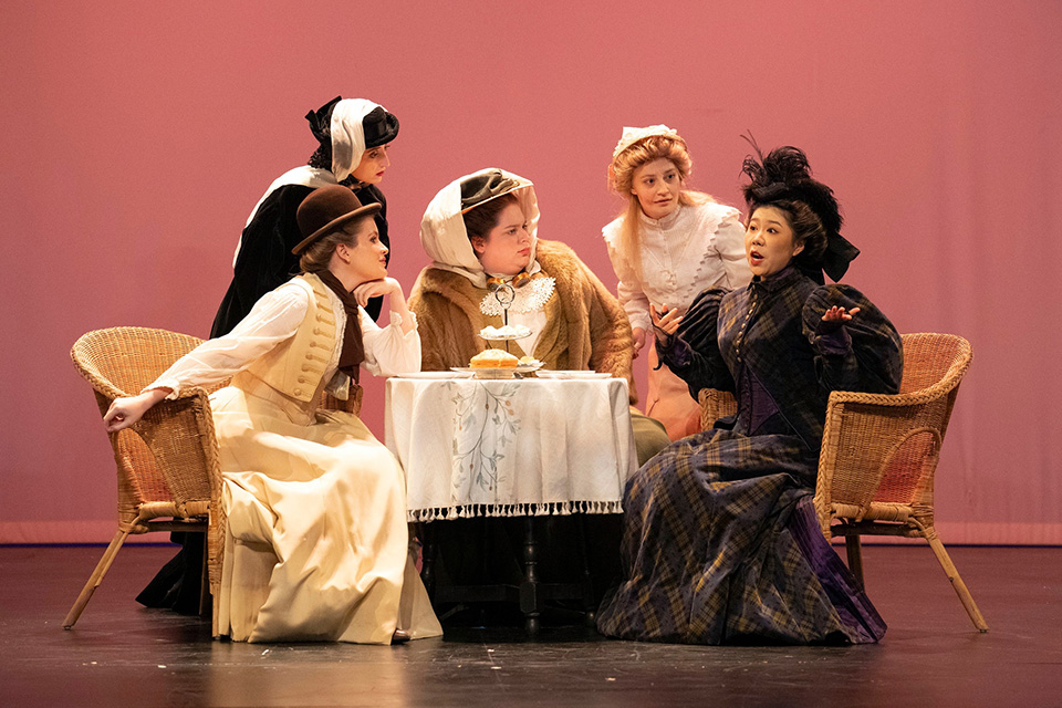 A group of opera singers in Victorian era clothing sit around a table mid-performance