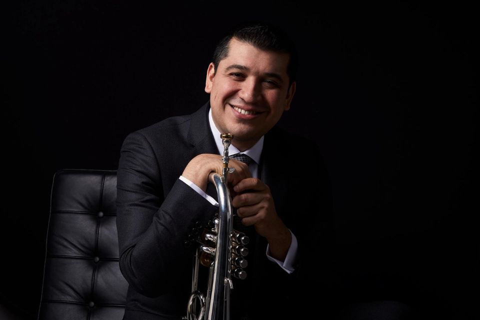 Pacho Flores wears a dark suit and holds his trumpet and smiles