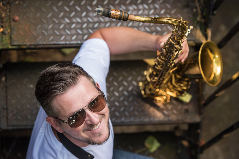 Paul Booth wears sunglasses and holds his saxophone, looking up at the camera smiling 