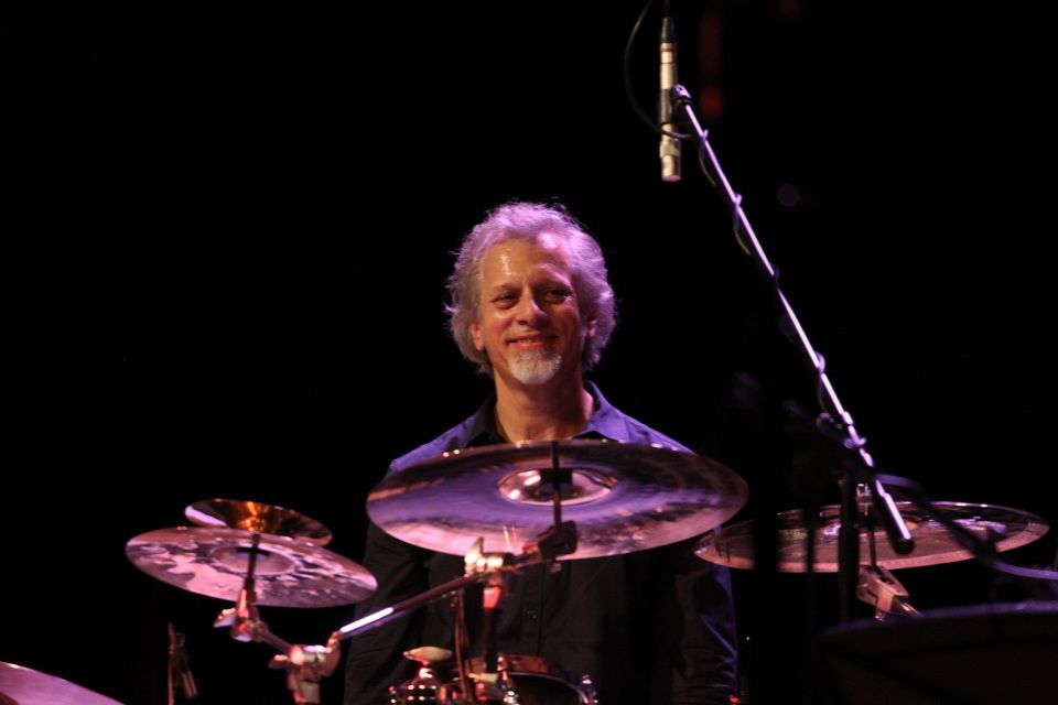 Dave Weckl sits behind a drum kit against a dark background and smiles