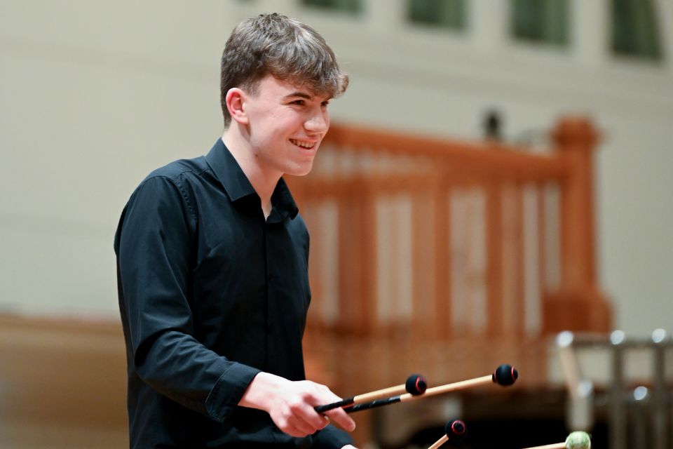 A young man wears a black shirt and holds percussion mallets, smiling