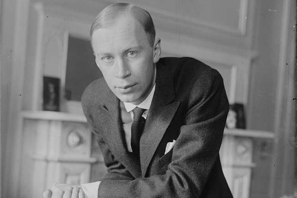 A black and white photo of Sergei Prokofiev wearing a suit and leaning on a chair back