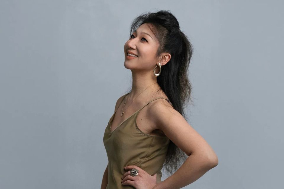 A young woman has a hand on her hip and smiles upwards against a grey background