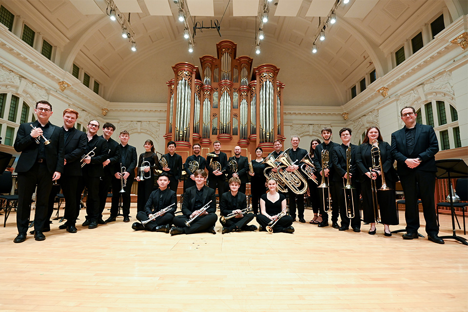 Members of the RCM Brass Ensemble stand and sit in two lines on stage, smiling at the camera