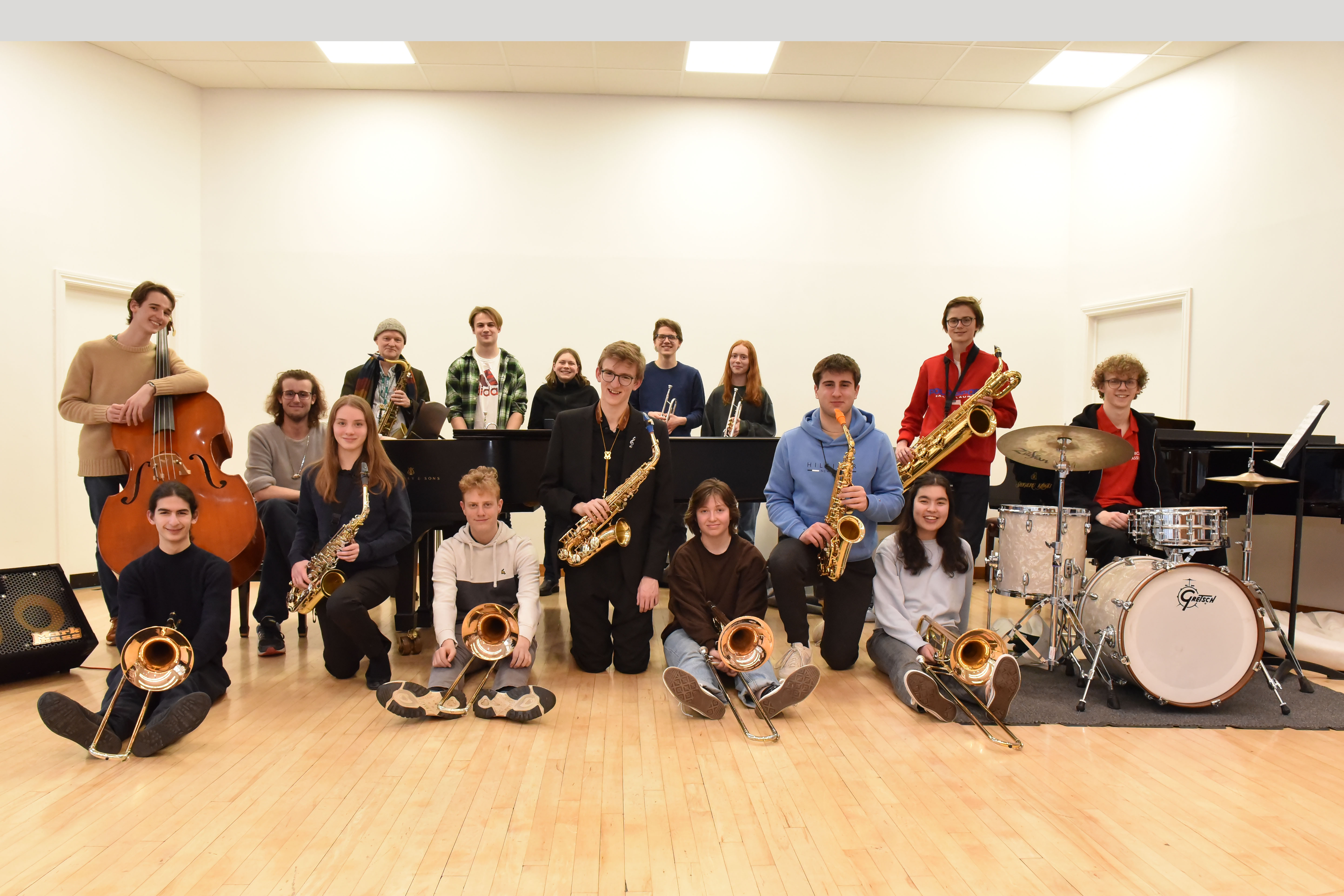 A group photograph of young people holding saxophones, trumpets and other jazz instruments, as well as someone sitting at a drum kit, in a room with bare white walls and a wooden floor