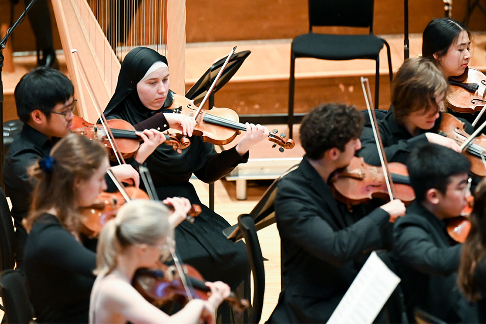 Violinists in a string orchestra perform on stage wearing all black