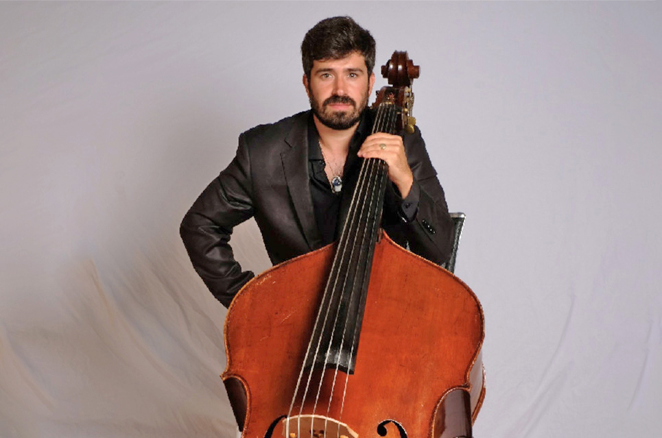 Rodrigo Moro Martin standing in front of a plain grey background wearing a black suit and holding his double bass